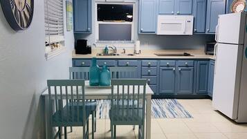 Une cuisine avec des meubles de couleur bleue, un réfrigérateur blanc, une horloge et une fenêtre avec des stores.