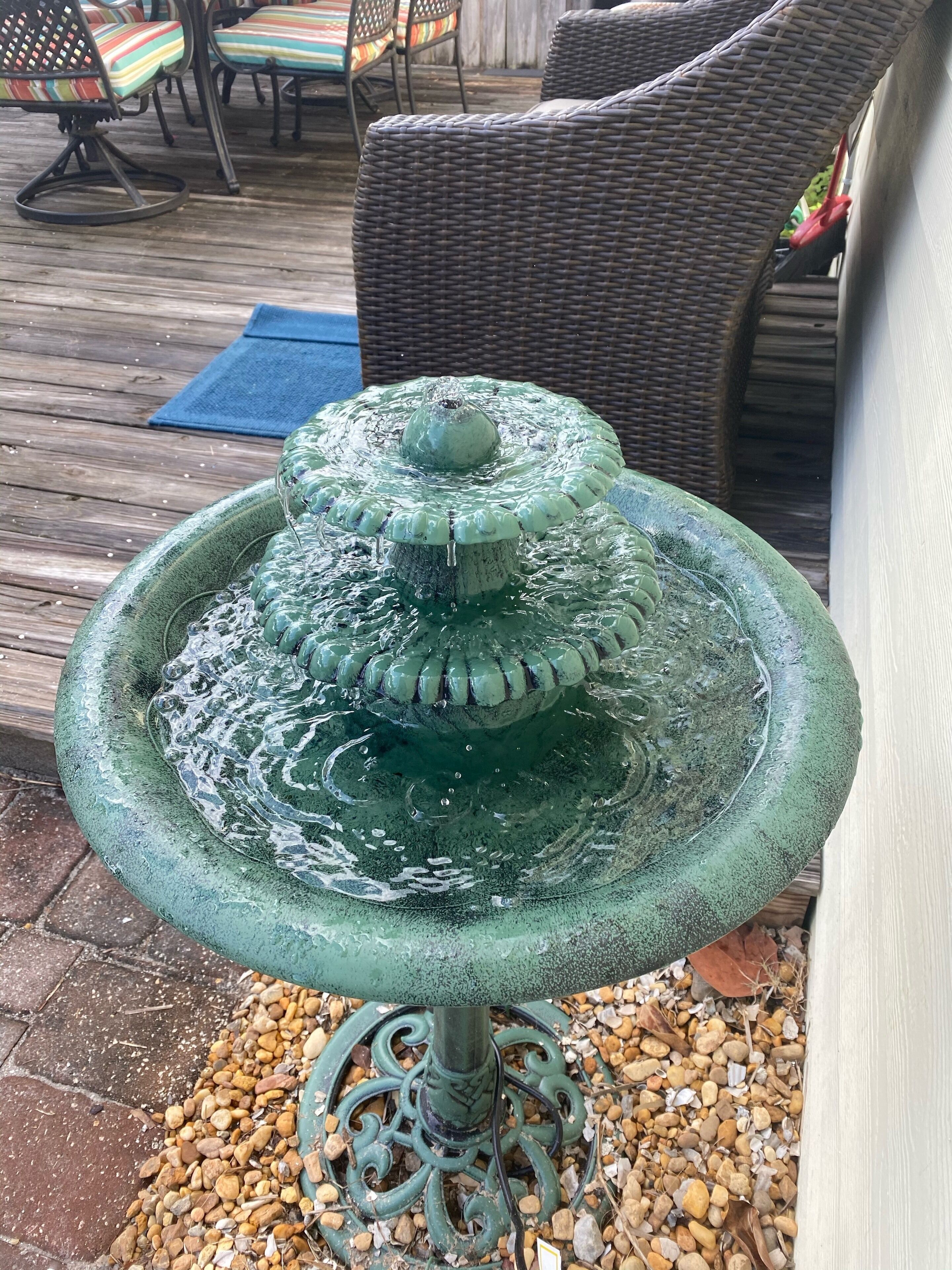 Eine grüne Außenbrunnen mit gestufter Bauweise, umgeben von Kieselsteinen und Korbmöbeln im Hintergrund.