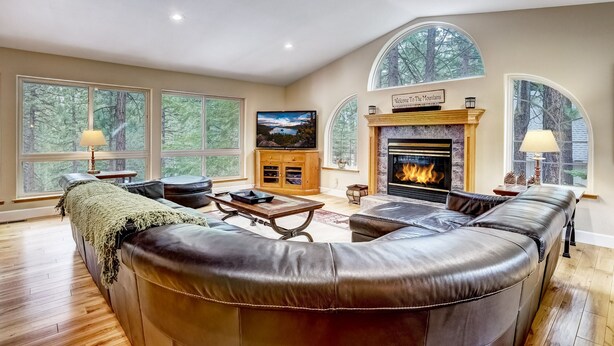 A spacious living room with a large leather sectional sofa, a fireplace, a television, and large windows offering a view of trees.