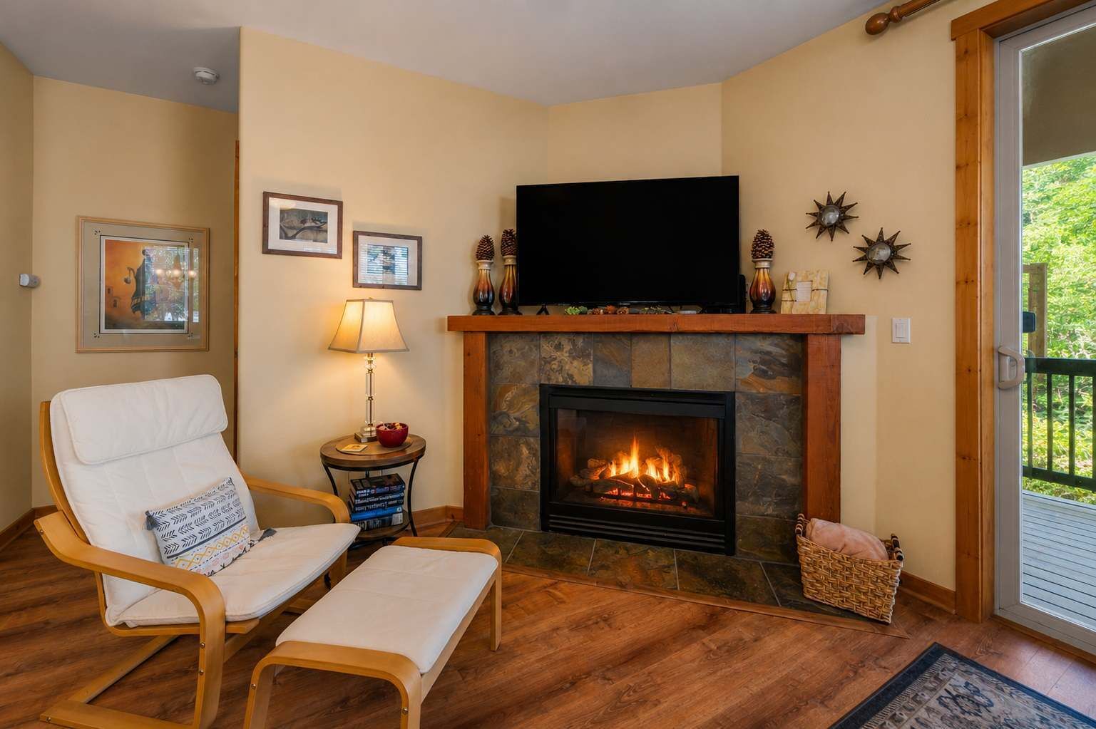 A living room with a fireplace, a television, a chair, a table with a lamp, and framed pictures on the wall.