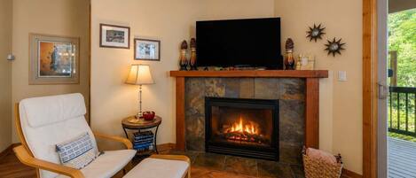 A living room with a fireplace, a television, a chair, a table with a lamp, and framed pictures on the wall.