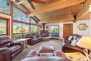 A spacious living room with leather sofas, a fireplace, large windows, and a wooden ceiling.