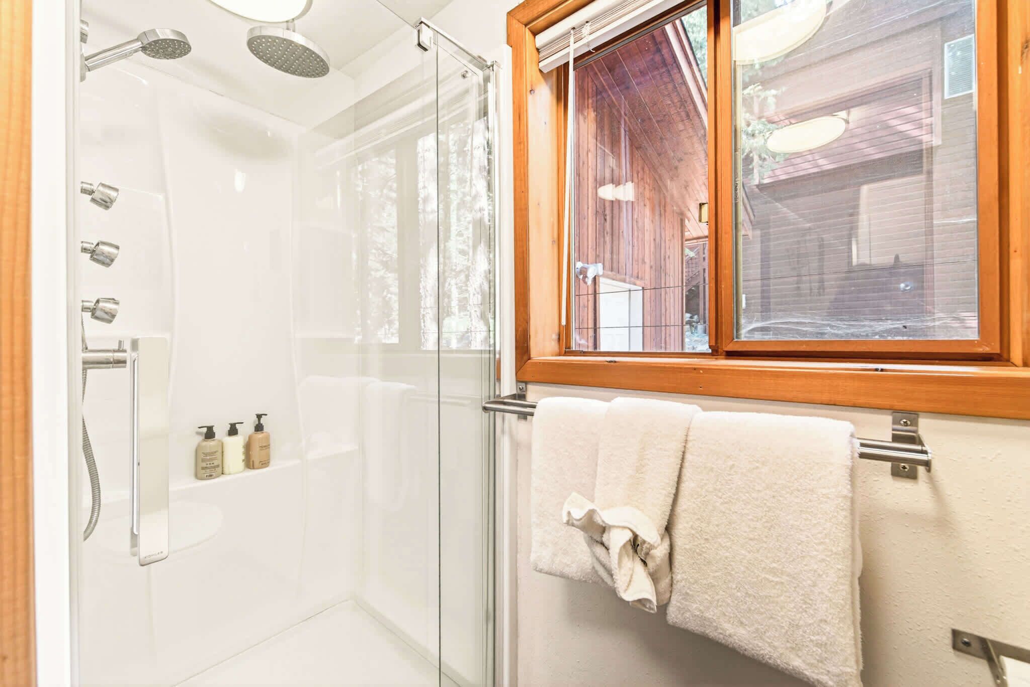 A glass-enclosed shower with multiple showerheads, a window with a wooden frame, and a towel rack with a towel.