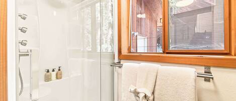 A glass-enclosed shower with multiple showerheads, a window with a wooden frame, and a towel rack with a towel.