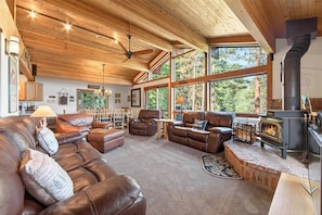 A cozy living room with leather sofas, a fireplace, and large windows overlooking a forest.
