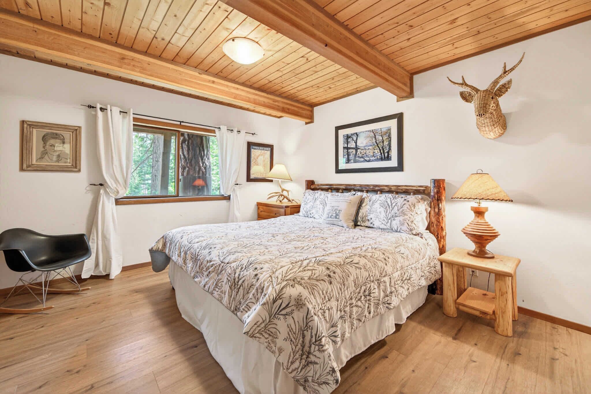 A bedroom with a wooden ceiling, a bed with patterned bedding, a rocking chair, bedside lamps, and a deer head decoration.