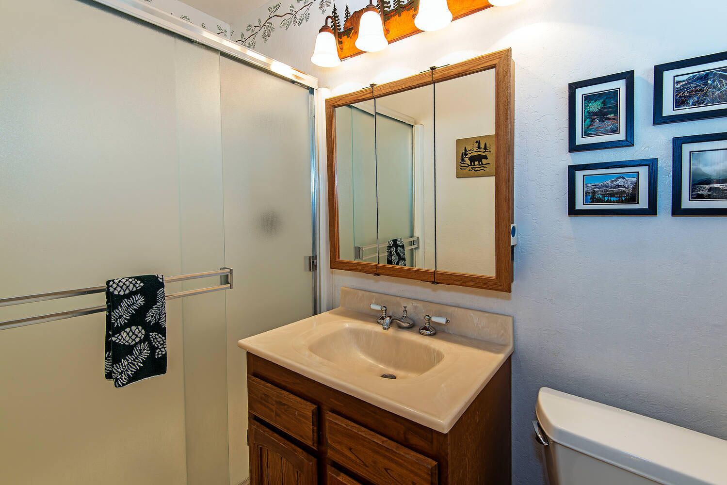 Une salle de bain avec un lavabo beige, un meuble en bois et un miroir au-dessus. Des tableaux encadrés ornent le mur et une serviette est suspendue à un porte-serviettes.