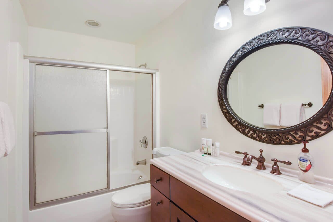 A bathroom with a white toilet, a glass shower enclosure, a white sink with a round mirror, and a wooden vanity.