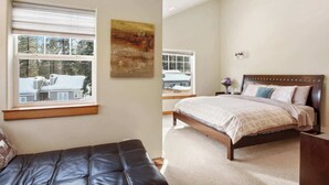 Une chambre à coucher avec un lit, une fenêtre avec des stores, un tableau au mur et une vue sur une maison à l’extérieur.