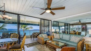 A spacious balcony with a glass wall offering a view of the ocean, furnished with a sofa, chairs, and a glass-top table. - Kona Reef #d31 Lanai w/ Coastal Views (Kailua-Kona)