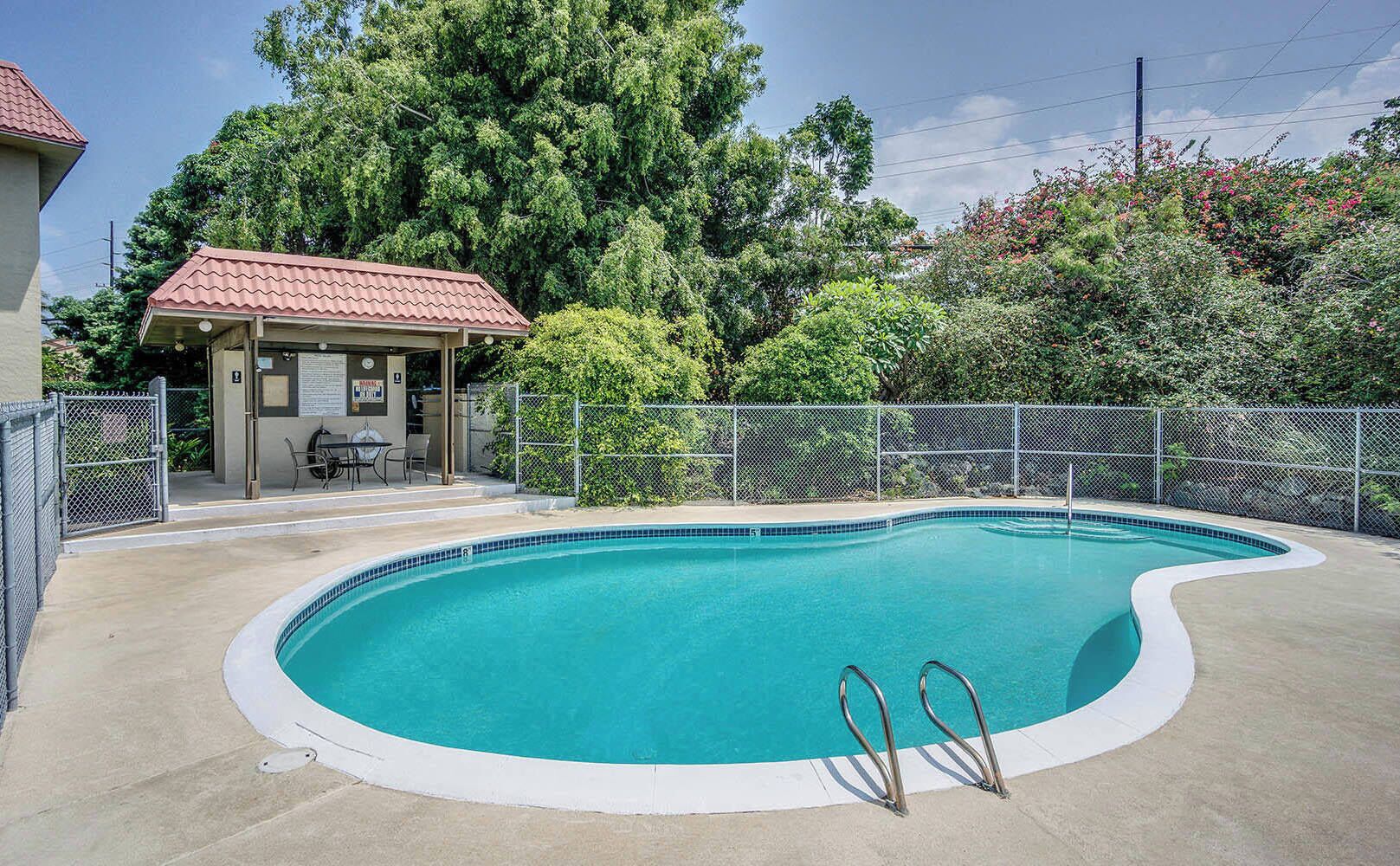 A swimming pool with a curved edge, surrounded by a fence and a covered seating area with a table and chairs.