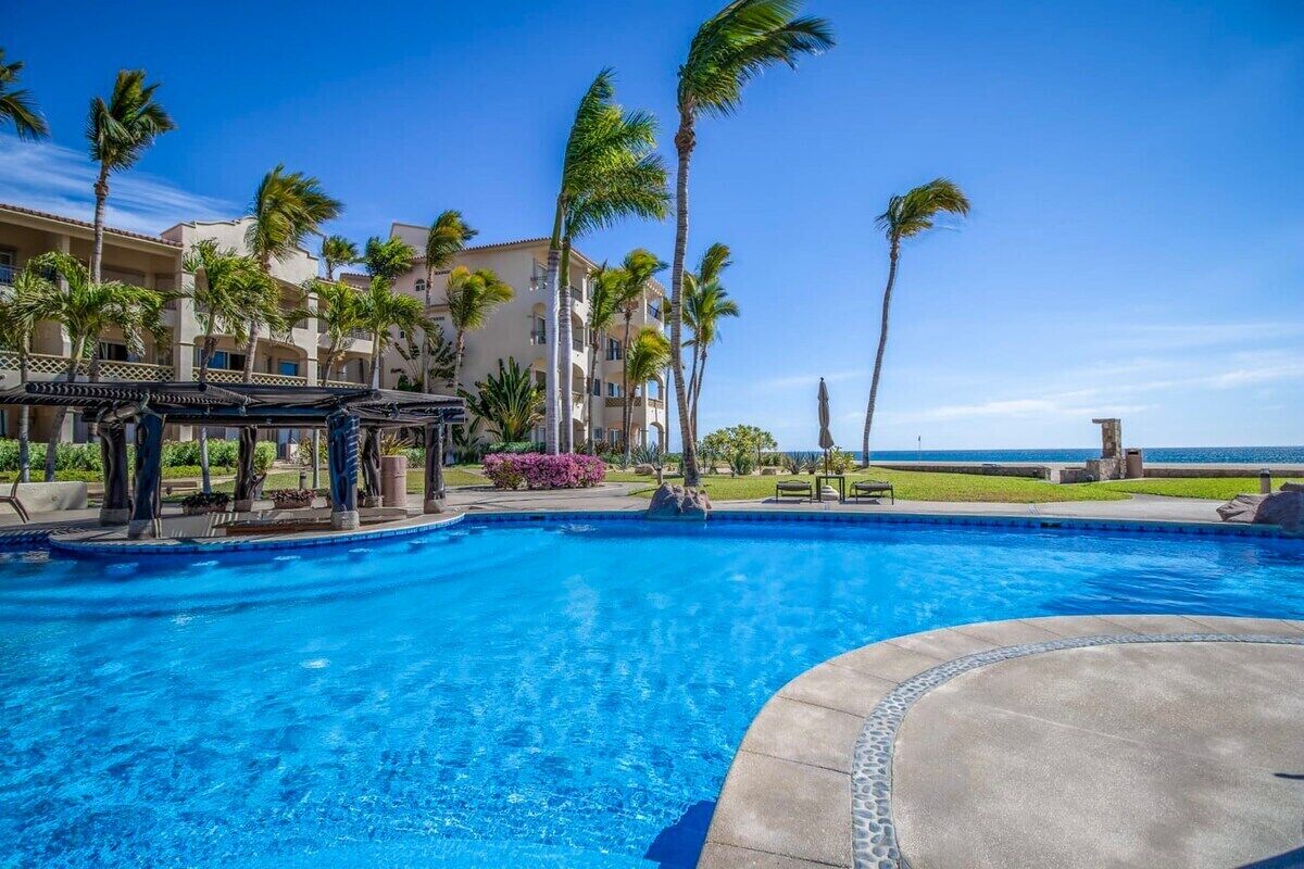 A swimming pool with clear blue water, surrounded by palm trees and a building with balconies.