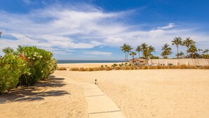 Una playa arenosa con cielo despejado, palmeras y un camino que lleva a un edificio.