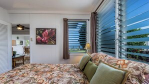 Un dormitorio con colcha estampada, vista a través de una ventana con persianas, un cuadro de una flor y una lámpara en la mesita de noche.