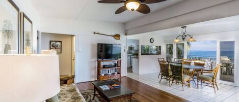 A spacious living area with a ceiling fan, a dining table with chairs, a television, and a view of the ocean.