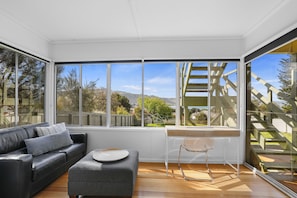 A modern living room with a leather sofa, a grey ottoman, and a clear glass table. Large windows offer a view of a wooden staircase and a mountainous landscape. - Sea Forever (Apollo Bay)