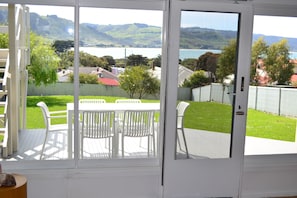 A view through a glass door of a patio area with a table and chairs, a wooden deck, and a fenced yard with a hilly landscape in the background.