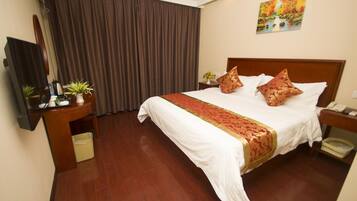 A hotel room with a large bed, a wooden headboard, a flat-screen TV, a desk with a plant, and a painting on the wall.