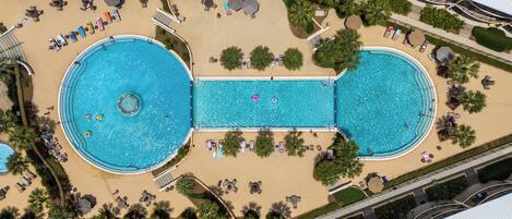 An aerial view of a resort with two large swimming pools, sun loungers, and palm trees.