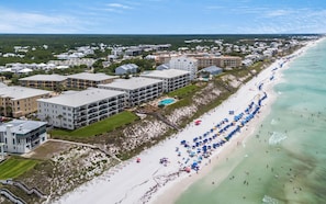 An aerial view of a coastal resort with multiple buildings, a sandy beach with umbrellas, and a clear view of the ocean.