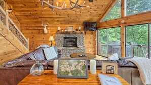 A cozy cabin interior with a stone fireplace, wooden walls, a leather sofa, and a large window offering a view of the outdoors. - Hot Tub, 3 Fireplaces, Mountain Views, Game Room (Blue Ridge)