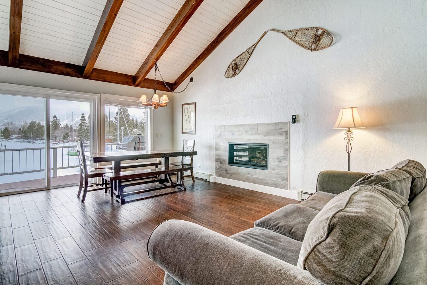A spacious living room with a wooden floor, a fireplace, a dining table with chairs, and a large window offering a view of the outdoors.