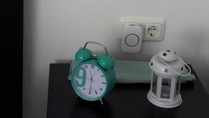 A small bedside table with a green alarm clock, a white lantern, and a wall-mounted desk lamp. - Studio Apartment , 33m2 (Bad Homburg vor der Höhe)