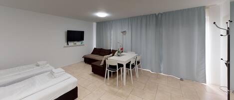 A hotel room with a bed, a sofa, a dining table with chairs, a TV mounted on the wall, and a window with curtains.