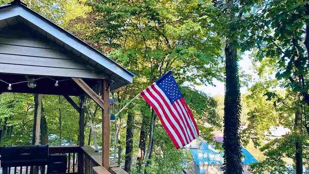 A wooden deck with a metal roof, an American flag, and a view of a lake with a building in the background.