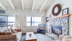 A living room with a stone fireplace, a television, and a view of the sea. - Waters Edge Vista HOA Pool & Pier Access (Tahoma)