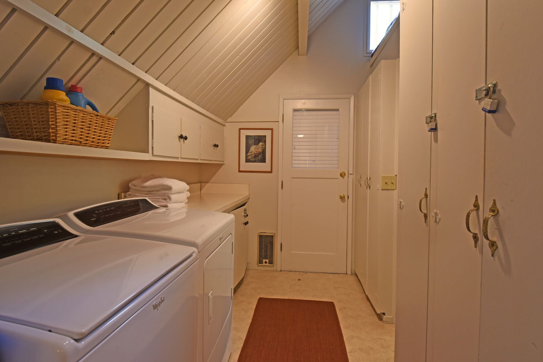 A laundry room with a front-loading washer and dryer, a wicker basket with bottles, a framed picture on the wall, and a door with a window.
