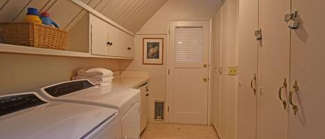 A laundry room with a front-loading washer and dryer, a wicker basket with bottles, a framed picture on the wall, and a door with a window.