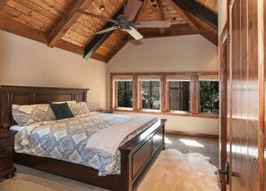 Une chambre avec un plafond en bois, un lit recouvert d’une literie à motifs, un ventilateur de plafond et de grandes fenêtres donnant sur la forêt.