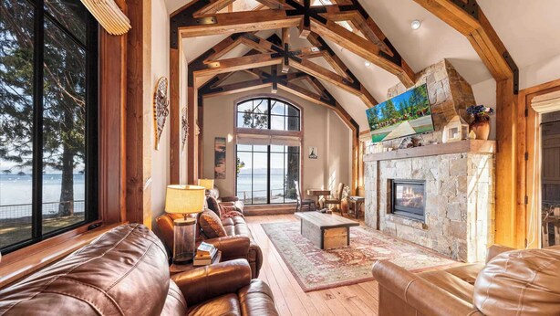 A spacious living room with a stone fireplace, leather sofas, and a large window overlooking a scenic view. - West Lake Serenity 180 Degree Lake Views (Homewood)