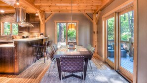 A spacious dining area with a long table and chairs, a kitchen with wooden beams, and large windows offering views of the outdoors. - Shining Shire - Sauna, Steam Shower, Very Cool! (South Lake Tahoe)