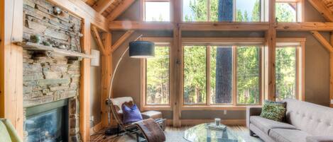 A cozy cabin interior with a stone fireplace, wooden beams, large windows, and a comfortable seating area.