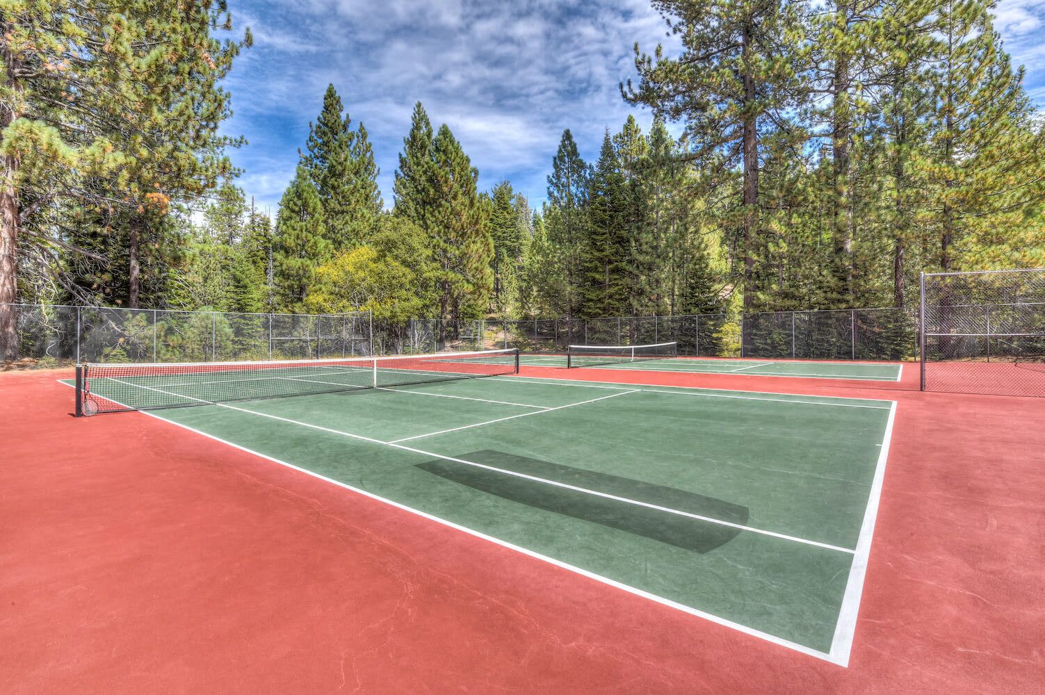 Cancha de tenis exterior con red, rodeada de árboles y un cielo despejado.
