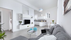A modern living room with a grey sofa, a glass coffee table, and a TV on a white stand. There is a kitchen in the background with white cabinets and a dining area. - Villa Anna with private Terrace and Sea View (Positano)