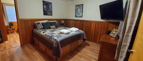 Une chambre à coucher avec un lit en bois, deux téléviseurs fixés au mur et une table de chevet avec une lampe.