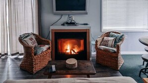 Un salon avec une cheminée, deux fauteuils en osier, un téléviseur et une fenêtre avec des stores.