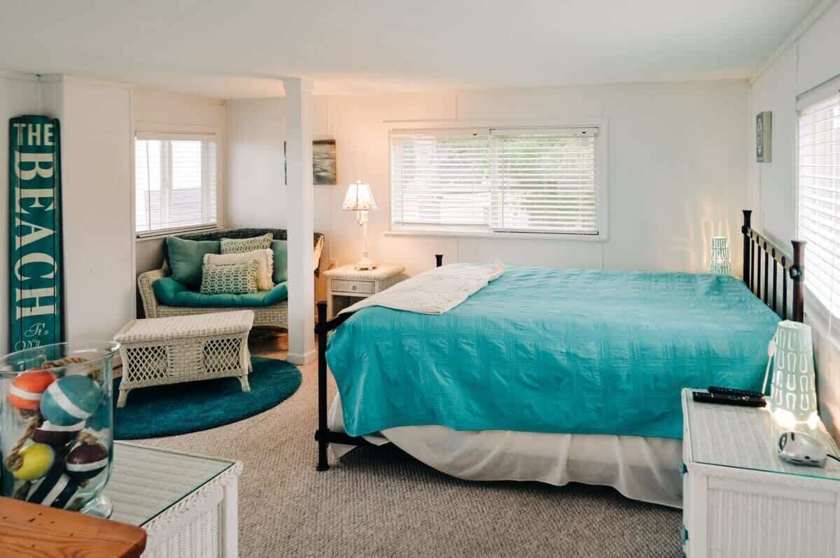 A bedroom with a bed, a nightstand, a lamp, a wicker chair, and a wall sign.