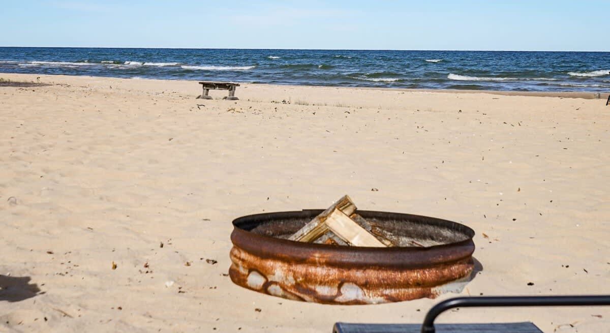 Una playa con un hogar a fuego de metal que contiene leña, un banco y el océano al fondo.