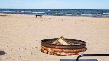 Una playa con un hogar a fuego de metal que contiene leña, un banco y el océano al fondo.
