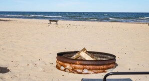 A beach with a metal fire pit containing logs, a bench, and the ocean in the background. - Charming Lake Huron Cottage Private Beach Access (Oscoda)