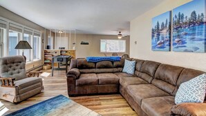 A living room with a brown leather sectional sofa, a grey armchair, a wooden coffee table, and a blue rug. There is a bookshelf, a dining table with chairs, and a ceiling fan.