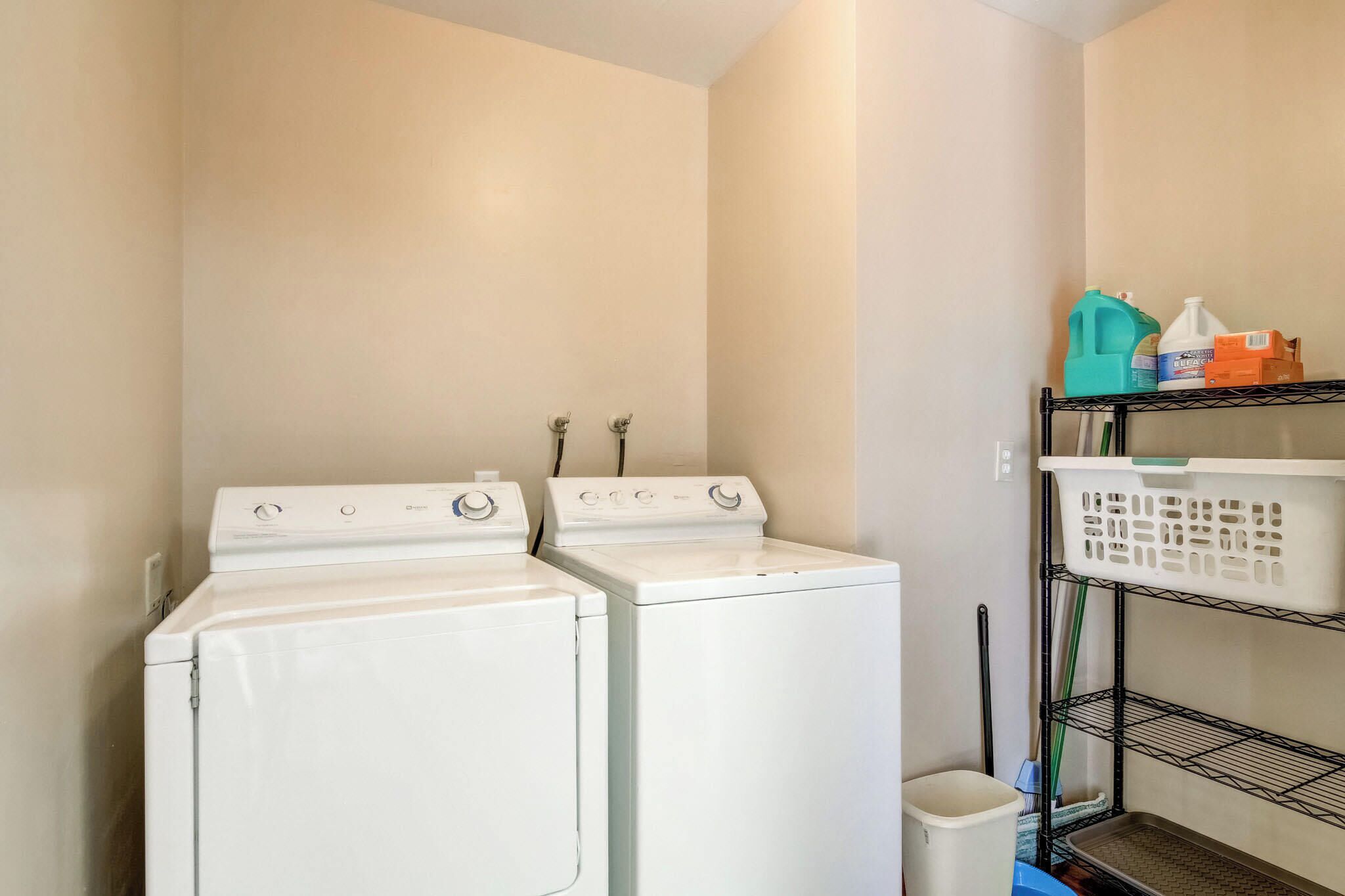 A laundry room with two white front-loading washing machines, a metal shelf with laundry baskets, and cleaning supplies.