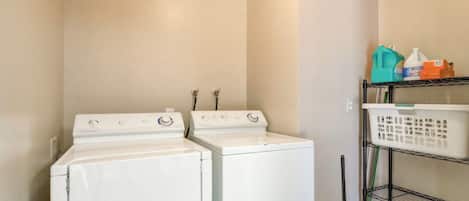 A laundry room with two white front-loading washing machines, a metal shelf with laundry baskets, and cleaning supplies.