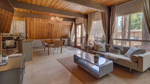 A modern interior with a wooden ceiling, a kitchen area, a living room with a sofa and coffee table, and large windows offering a view of trees and buildings.