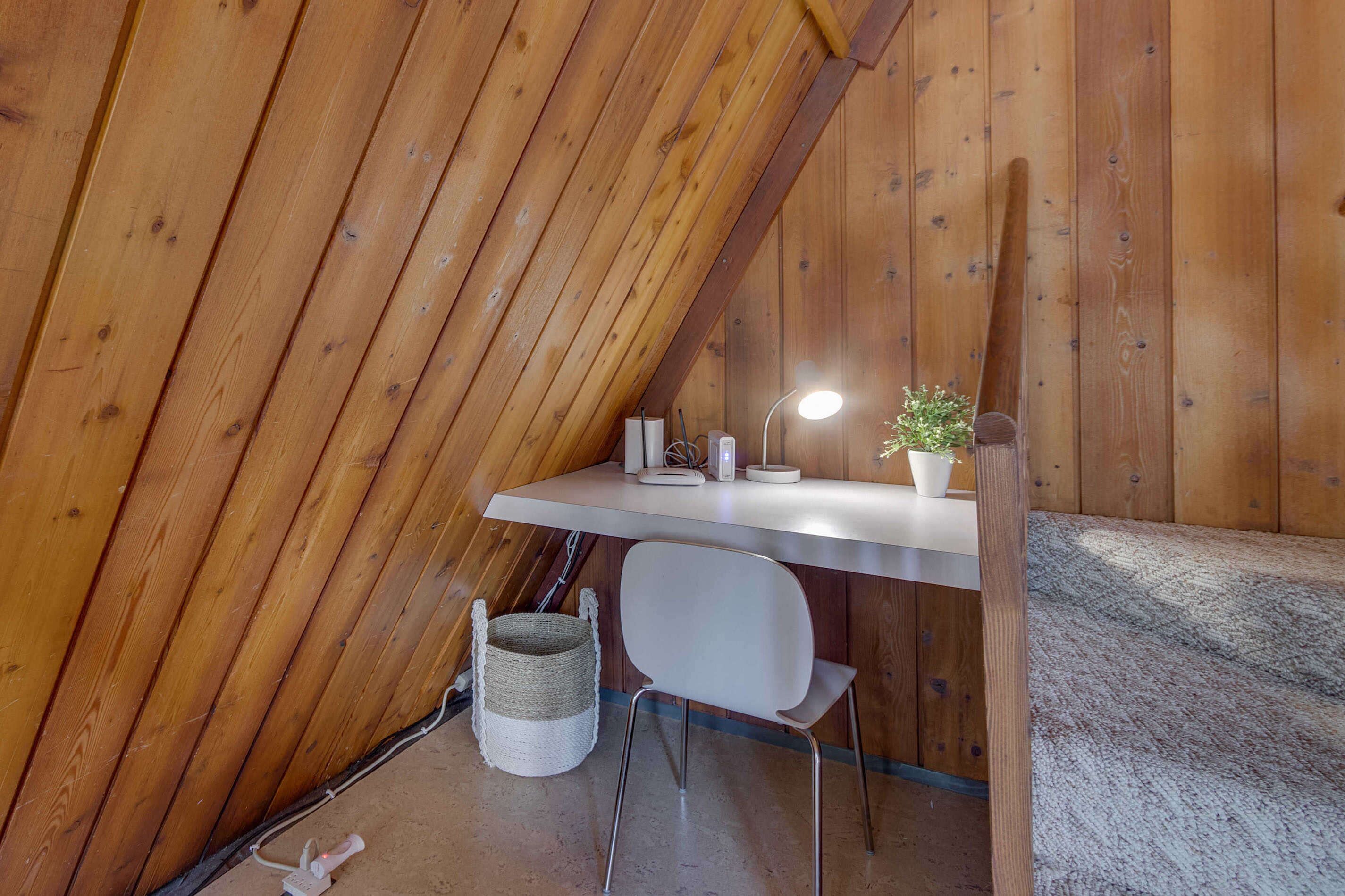 A small, cozy attic bedroom with a slanted ceiling, a white desk with a lamp, a chair, and a small potted plant.