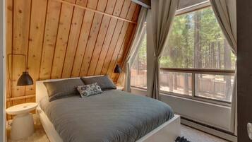 Un dormitorio con techo inclinado, una cama grande, dos lámparas de noche, una mesita pequeña y vistas a árboles a través de una ventana.